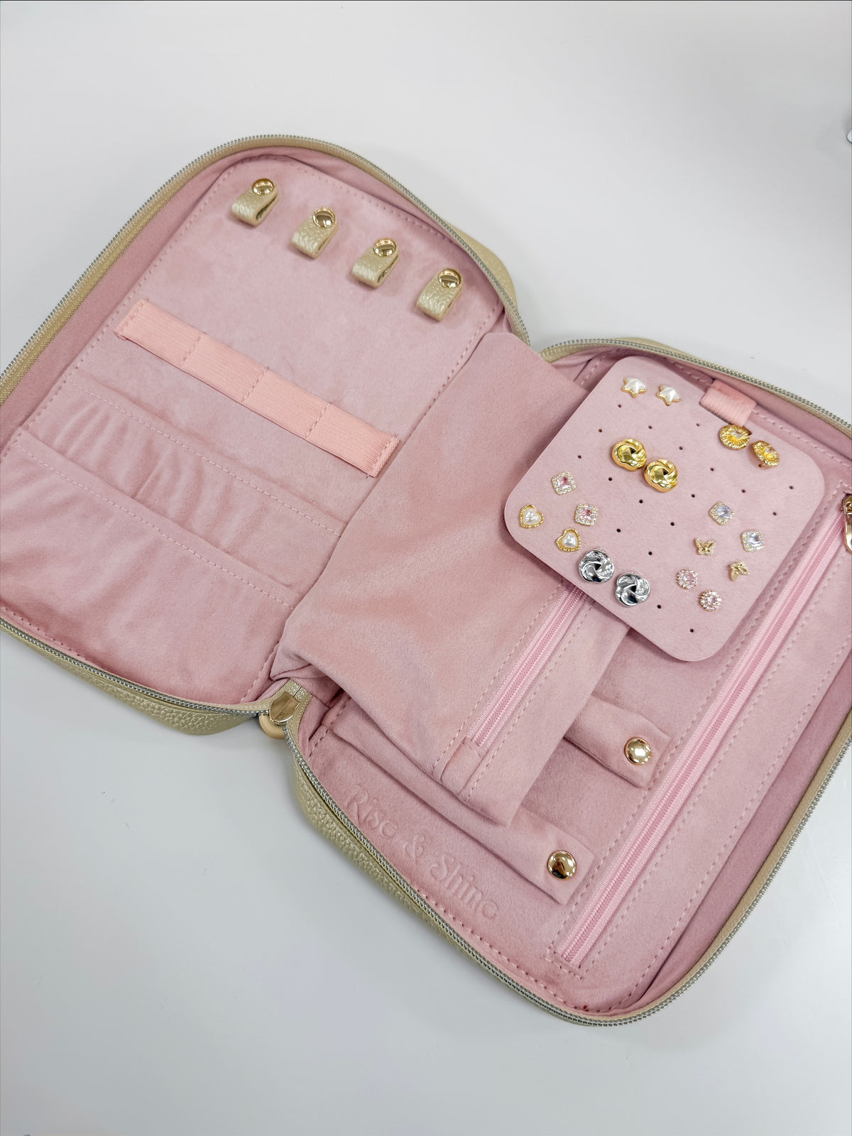 An open pink jewelry organizer case with gold-tone zippers and hardware, showing interior slots, a small matching pocket with rhinestone stud earrings, and a pearl necklace displayed inside.