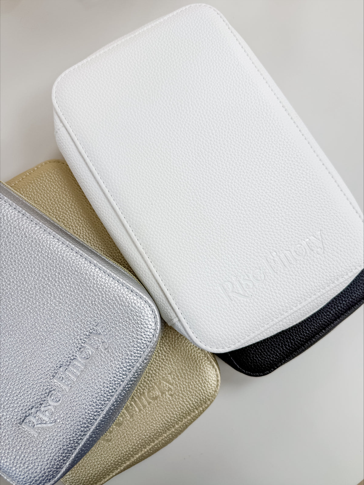 Three textured leather zippered pouches stacked diagonally on a light neutral background. The top pouch is white, the bottom-left is metallic silver, the bottom-center is metallic gold, and a black edge is visible behind the gold pouch.