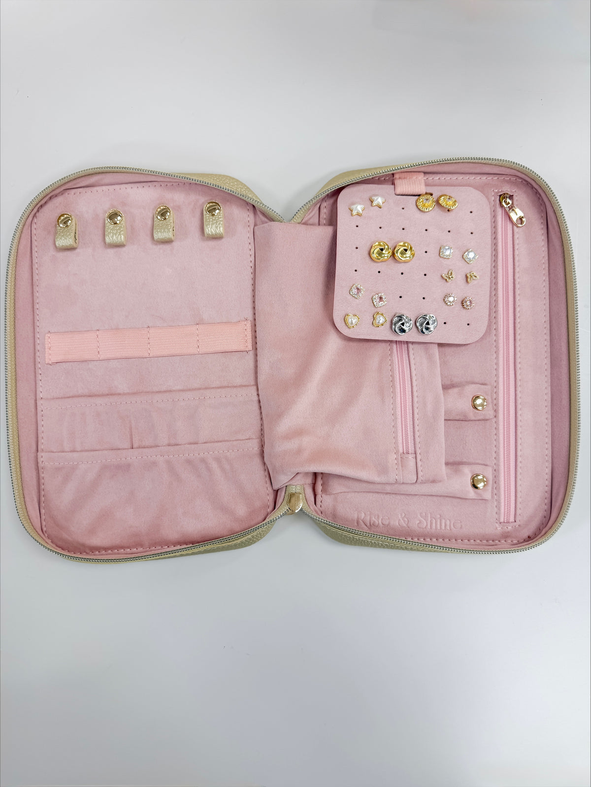 An open pink jewelry organizer case with a light beige exterior trim, featuring multiple interior slots, a small pocket with decorative studs and crystals, and gold-tone rings on the left side.