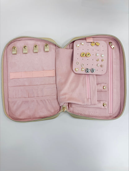 An open pink jewelry organizer case with a light beige exterior trim, featuring multiple interior slots, a small pocket with decorative studs and crystals, and gold-tone rings on the left side.